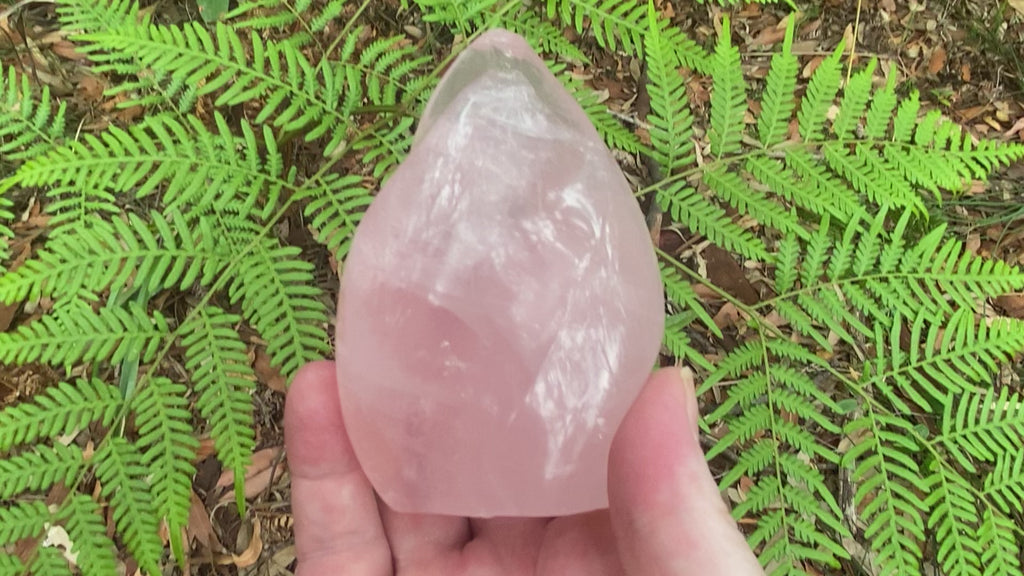 Video shows a polished rose quartz crystal in the shape of a flame. H 10 cm W 6.5cm D 6.5cm